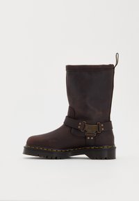 Brown leather ankle boot featuring a side strap with metallic accents, a textured surface, and a thick, ridged rubber sole with yellow stitching.