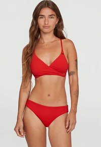 Sieviete ar gariem brūniem matiem, tērpta sarkanā peldkostīmā, stāv pret vienkāršu gaišu fonu, rādīdama tetovējumus uz rokām.