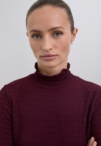 Burgundy long-sleeve top with a textured fabric, high ruffled collar, and tight fit, featuring a smooth finish and subtle accents.