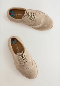 Chaussures brogue en daim beige avec un bout rond, perforations décoratives et lacets. Tige texturée et semelle contrastante. La doublure intérieure arbore un design coloré.