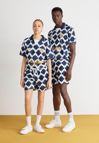 Marineblaues und weißes gemustertes Sportoutfit aus zwei Teilen, bestehend aus einem Kragen, kurzen Ärmeln und passenden Shorts mit einem Kordelzug. Goldenes Logoakzent.