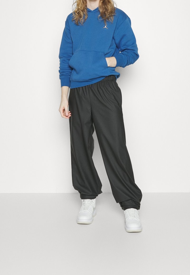 Osoba v modré mikině s malým logem, černých volných kalhotách a bílých teniskách stojí na pozadí s jednoduchou bílou barvou.