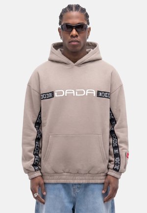 Man with braided hair and sunglasses wearing a beige DADA hoodie with black side stripes and blue jeans, standing against a plain background.