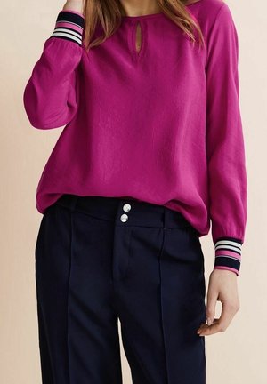 Frau trägt eine langärmelige magentafarbene Bluse mit gestreiften Manschetten und marineblaue, hoch taillierte Hose mit doppeltem Knopfverschluss.