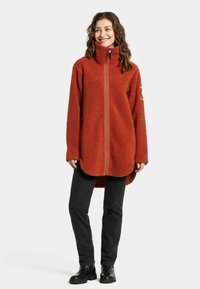 Fleecejacka i bränd orange med hög krage, fullständig frontblixtlås, långa ärmar och rundad nederkant. Svarta byxor och stövlar fulländar outfiten.