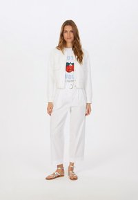 Weißer strukturierter Blazer über einem Grafik-T-Shirt mit Erdbeerdesign, kombiniert mit weißen plissierten Hosen und silbernen Riemchensandalen.