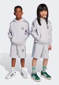 Ljusgrå huvtröjor har framfickor och broderade logotyper. Matchande grå shorts med sidostreck. En modell bär gröna sneakers, den andra vita.