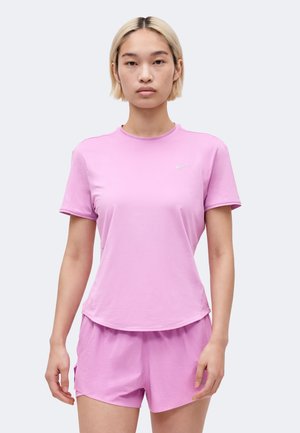 Giovane con capelli biondi corti che indossa una t-shirt sportiva Nike rosa chiaro e pantaloncini abbinati, in piedi rivolto verso di fronte.