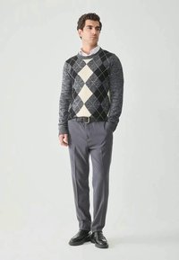 Man standing, wearing gray argyle sweater, white collared shirt, gray trousers, black belt, and black shoes against plain light background.