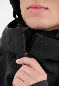 Svart jacka med en slät textur, med dragkedjestängning och perforerade detaljer. Kragen är hög och en hand greppar dragkedjehandtaget.