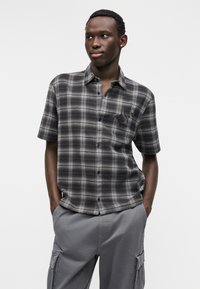 Κοντομάνικο πουκάμισο με καρό σε σκουρό γκρι και μπεζ ύφασμα με λεπτομέρεια τσέπης. Φορεμένο με γκρι παντελόνι cargo, χέρια στις τσέπες.