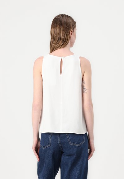 Weißes ärmelloses Top mit einem Schlüsselrücken-Detail, aus glattem Stoff gefertigt. Kombiniert mit dunkelblauen High-Waist-Jeans.