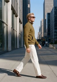 Homme portant des lunettes de soleil, une veste verte texturée, un pantalon blanc et des chaussures marron marchant sur un trottoir en ville entre de hauts immeubles en plein jour.