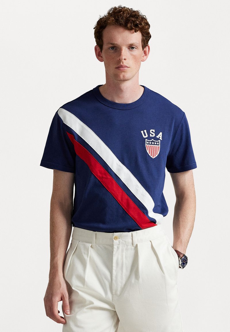 Tricou din bumbac Navy cu dungi diagonale roșii și albe. „USA” brodat pe piept cu un logo în formă de scut. Design cu mâneci scurte și guler rotund.