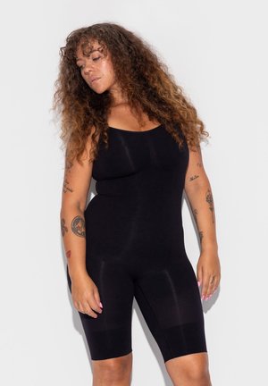 Vrouw met lang krullend haar die een zwarte mouwloze body draagt, met tatoeages op beide armen, staande tegen een witte achtergrond.