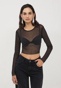 Top lungo a maniche nere in mesh con pois bianchi e design cropped, indossato con un reggiseno nero sotto e abbinato a jeans neri a vita alta.