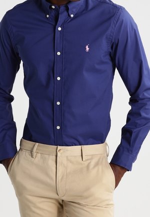 Homme portant une chemise bleu marine ajustée à boutons avec un logo brodé rose, associée à un pantalon beige, les mains dans les poches.