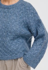 Sininen neulepaita, jossa on kaapelikuosi, pyöreä pääntie ja pudotetut olkapäät. Valmistettu pehmeästä, teksturoidusta langasta ja lyhyestä istuvuudesta.