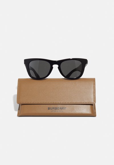 Fekete macskaszem napszemüveg sötét lencsékkel egy bézs bőrtok tetején, amelyen fémes betűkkel a "BURBERRY" felirat látható.