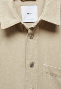 Camisa de botones beige hecha de tela texturizada que cuenta con un cuello, botones plateados y un solo bolsillo en el pecho. La etiqueta es visible cerca del cuello.