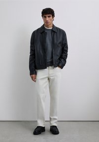 Hombre con chaqueta de cuero negra sobre suéter gris, pantalones blancos y zapatos negros, de pie contra una pared simple de color gris claro.