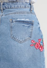 Shorts en denim bleu avec une poche arrière usée brodée de fleurs rouges et bleues, présentant un tissu texturé et un design décontracté.