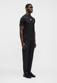 Μαύρο κοντομάνικο μπλουζάκι με λογότυπο στο στήθος, σε συνδυασμό με μαύρο παντελόνι στενής γραμμής και μαύρα αθλητικά παπούτσια, παρουσιάζεται σε απλό λευκό φόντο.