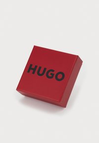 Scatola rettangolare rossa con il testo nero in evidenza che scrive "HUGO." Superficie liscia e lucida con angoli e bordi affilati.