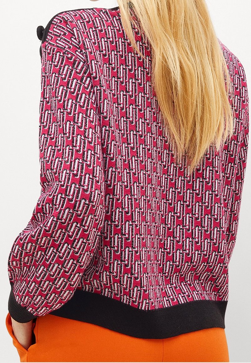 Femme aux longs cheveux blonds portant un pull rouge à motifs avec des poignets noirs et un pantalon orange, debout les mains dans les poches.