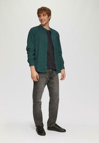 Teal button-down shirt met subtiel patroon, zwart t-shirt eronder, zwarte jeans met een scheur, en zwarte sneakers. Effen achtergrond.