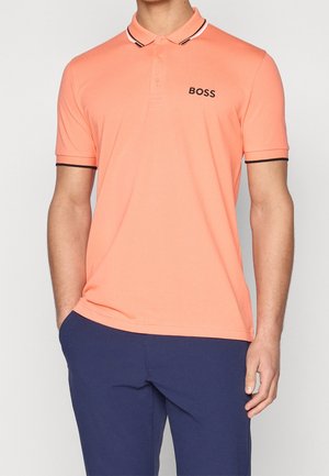 Muž nosící lososově růžové polo tričko s černým logem "BOSS" na hrudi a tmavě modré kalhoty, stojící s jednou rukou v kapse.