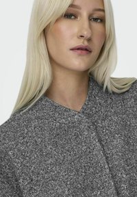 Grå, texturerad klädesplagg med V-halsdesign, som har ett mjukt, fluffigt material och en minimalistisk stil, sett bäras av en modell.