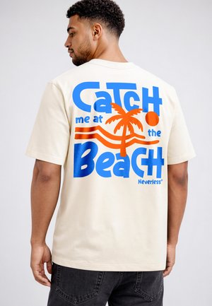 Man with short curly hair wearing a cream T-shirt with blue and orange text and palm tree graphic on back reading "Catch me at the Beach."