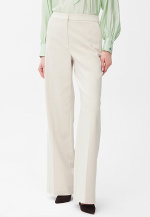 Vrouw draagt hoge taille, wijde beige broek gecombineerd met een lichtgroene blouse met lange mouwen en donkere schoenen met spits toelopende neus.