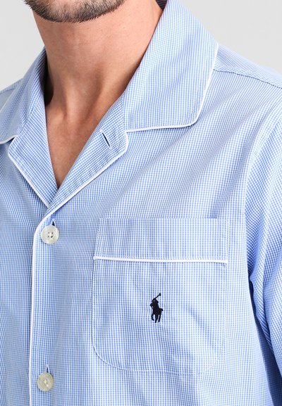 Camisa de pijama em padrão gingham azul e branco, com gola de bico, detalhes em branco e um único bolso no peito que apresenta um logótipo preto. Frente com fecho de botões.