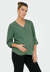 Blouse verte texturée, à col en V, avec des manches trois-quarts, fermeture à boutons et coupe détendue, associée à un pantalon noir.