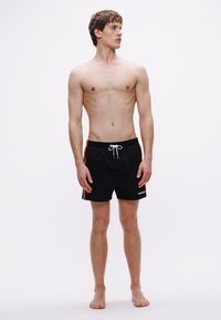 Pantaloni scurți de baie negri, cu talie ajustabilă cu șnur și dungi portocalii pe părți, realizați dintr-un material ușor și care se usucă rapid.