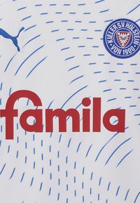 Balta sporta krekls ar zilu viļņu rakstu, lieliem sarkaniem uzrakstiem "famila" un apļveida zilā komandas logo ar ģerboni.