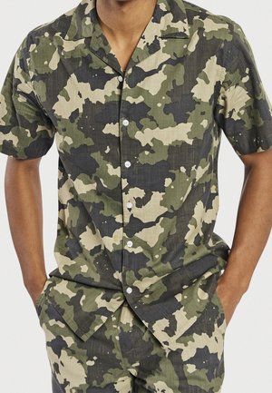 Mand iført grøn, beige og sort camouflaged kortærmet skjorte med knapper og matchende bukser med hænderne i lommerne.