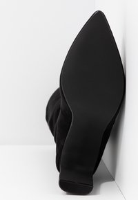 Vue unique d'une botte noire à talon et bout pointu, posée contre un coin blanc en arrière-plan.