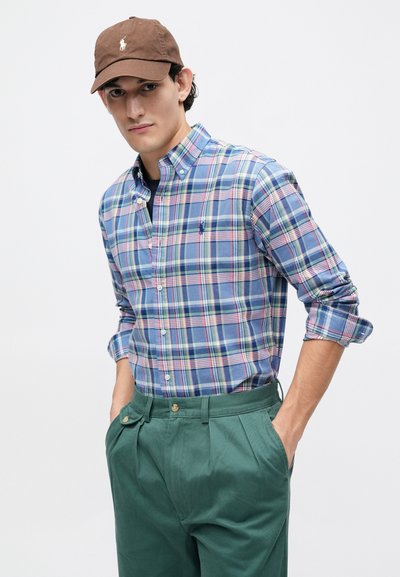 Πουκάμισο καρό σε μπλε, πράσινο και ροζ με γιακά κουμπί; πράσινα παντελόνια με πιέτες και κουμπιά; καπέλο καφέ με λογότυπο.