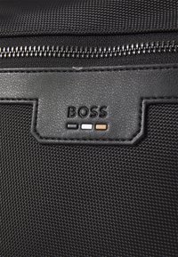 Juodas tekstūruotas audinio krepšys su ryškiu „BOSS“ logotipu, kuriame yra trys horizontalių akcentų spalvos: juoda, balta ir ruda.