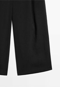 Pantalon noir à jambes larges et raccourci, montré de la cuisse vers le bas sur un fond blanc avec des détails visibles de la couture latérale.