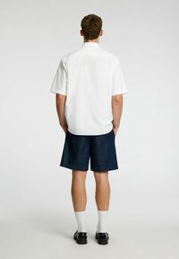 Camicia bianca a maniche corte, pantaloni corti di jeans blu scuro, calzini bianchi e scarpe nere. La vista posteriore mette in evidenza il collo della camicia e la lunghezza dei pantaloni.