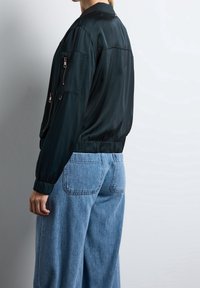 Sort satin bomberjakke med lynlåse foran, ribstrikkede manchetter og kant. Sat sammen med lyseblå jeans med vide ben. Glat tekstur, minimalistisk design.