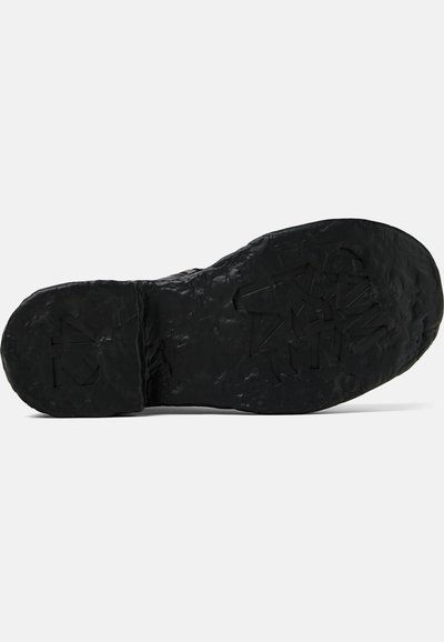 Suela de zapato negra con material de goma texturizada. Presenta una superficie rugosa e irregular y letras en relieve en la parte inferior para mejorar la tracción.