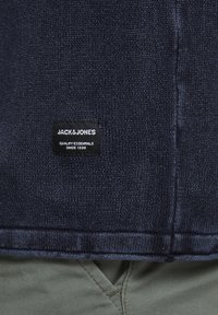 Pull en tricot bleu foncé avec une étiquette noire portant le texte blanc "JACK & JONES" et un ourlet texturé sur un tissu vert clair.