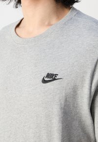 Šedá bavlněná mikina se širokým výstřihem. Obsahuje černě vyšívané logo Nike na levé hrudi. Měkká textura a ležérní design.