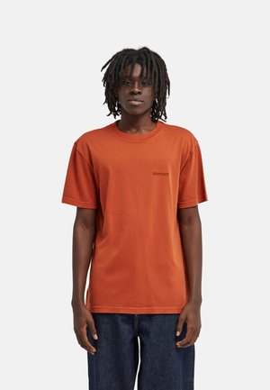 Person mit Dreadlocks, die ein oranges Kurzarm-T-Shirt mit dem "element"-Logo und dunkelblaue Jeans vor einem schlichten hellen Hintergrund trägt.