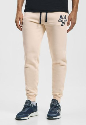 Mann trägt beige Joggerhosen mit marineblauen Kordeln und marineblauen Sneakers mit weißen Sohlen vor hellem Hintergrund.
